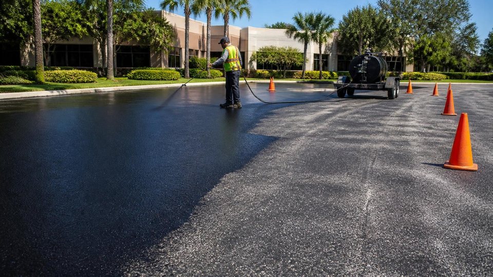 Worker spraying black sealer across an office park parking lot behind traffic cones